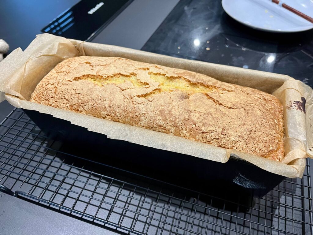 The image shows a freshly baked loaf of cake in a black rectangular baking pan, lined with parchment paper. The bread has a golden-brown crust and is sitting on a black wire cooling rack. The background includes a glossy black surface, possibly a stovetop, and out-of-focus tableware.