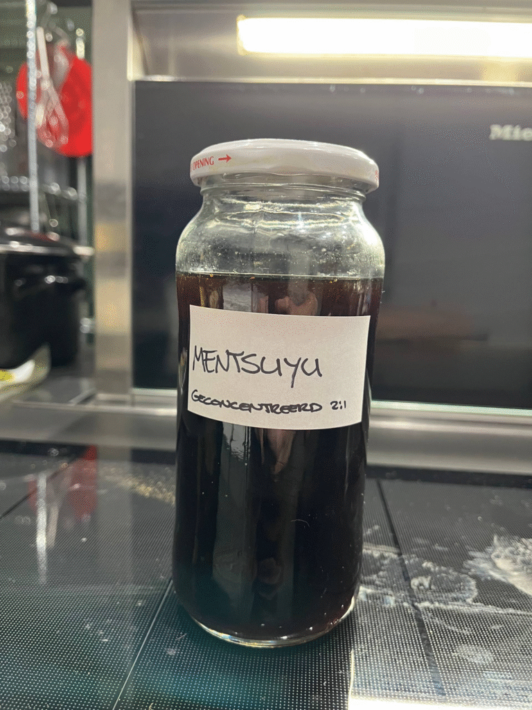 The image shows a glass jar filled with a dark liquid, labeled "Mentsuyu Geconcentreerd 2:1." The jar has a white lid with a red arrow and the word "OPENING" on it. The jar is sitting on a black, textured surface, likely a stovetop. In the background, there's a blurred view of kitchen utensils hanging on a rack and a Miele appliance.